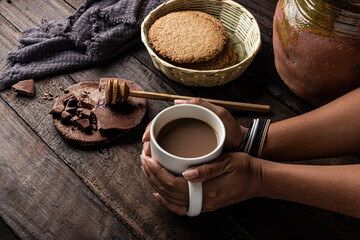 Manos de mujer sosteniendo taza de chocolate artesanal sobre mesa de madera antigua con galletas...