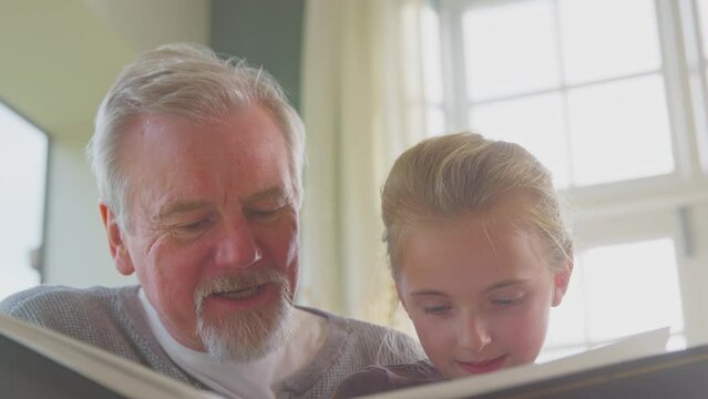 Close up of grandfather with granddaughter sitting in chair at home together looking through family photo album - shot in slow motion