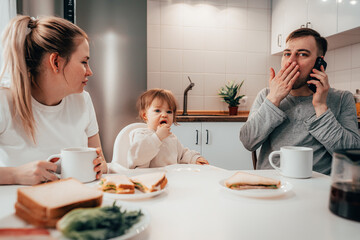 Happy smiling family is enjoying breakfast while father talk by cellphone in a kitchen at home in the morning.
