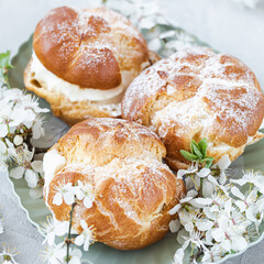 Choux Buns with whipped cream and sugar powder on top on plate. Gray background with spring white cherry blossom flowers. Delicate choux pastry dessert. French cream puffs, close-up