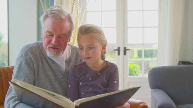 Grandfather with granddaughter sitting in chair at home together looking through family photo album - shot in slow motion