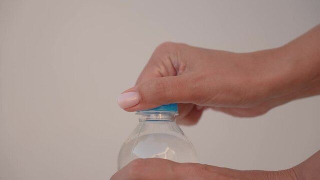 Close-up view 4k stock video footage of 500 ml transparent plastic bottle full of fresh water closed with blue plastic cap isolated on grey wall background. Woman opens bottle, water splashing out