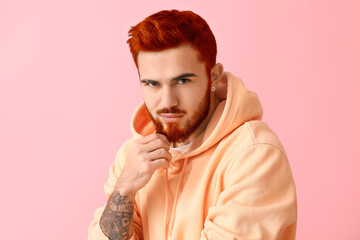 Stylish tattooed young man with unusual red hair and beard on pink background