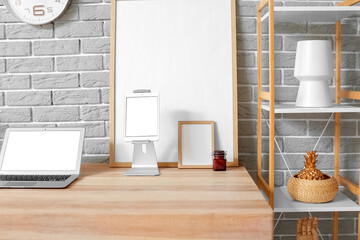 Modern laptop, tablet computer, blank photo frames on wooden standing desk and shelf unit near grey brick wall