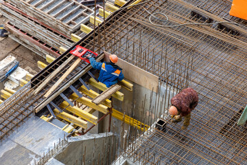 Builders in orange helmets make a steel reinforcement cage for pouring a concrete slab. Steel rods and metal formwork on a construction site. Monolithic construction. Cast-in-place concrete - obrazy, fototapety, plakaty