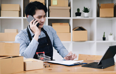 Young asia seller man happy work on laptop busy cellphone call chat to customer at home office desk...