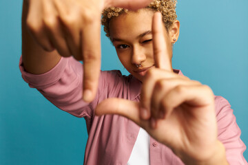 Selective focus of smiling black girl with blonde curly hair and piercing making frame with hands...