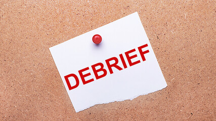 White paper with the text DEBRIEF is attached to the wooden background with a red button.