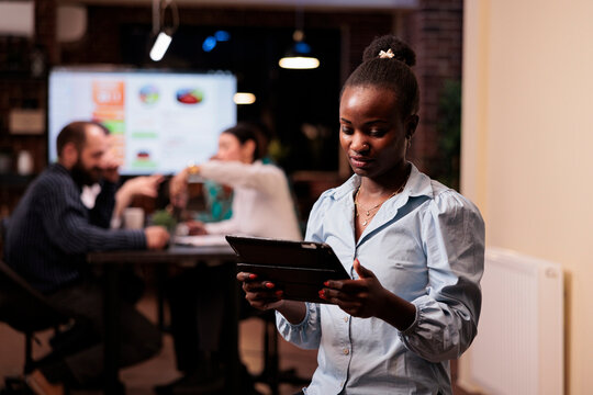 Focused african american woman holding tablet and looking at screen working late hours during business strategy meeting with mixed team. Startup employee posing confident in group project with