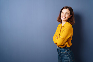 Laughing young woman stands with crossed arms in front of blue background and laughs into camera