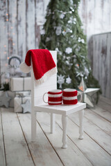Obraz premium Two mugs in knitted covers with a Santa hat are lying on a white wooden chair, Christmas background.