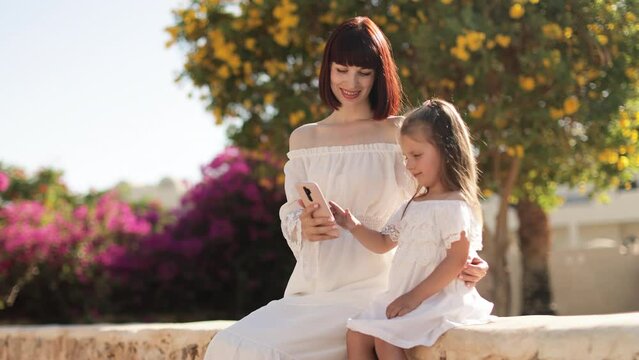 Portrait of good looking mother taking photo with daughter on mobile phone while sitting in the tropical park during summer vacation. Happy mom with daughter use mobile phone while sitting on stone.