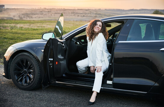 Beautiful businesswoman in elegant white suit sitting in modern electric vehicle. Charming young woman closing eyes with pleasure and smiling while posing in electro car.