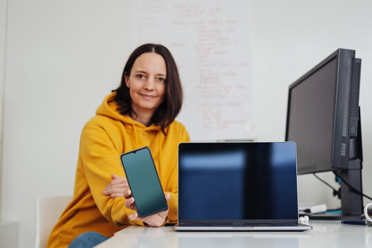 woman in office shows her cell phone and laptop screen to camera, copy space