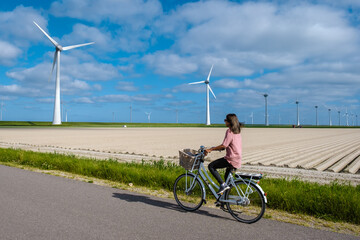 young woman electric green bike bicycle by windmill farm, windmills isolated on a beautiful bright day Netherlands Flevoland Noordoostpolder - obrazy, fototapety, plakaty