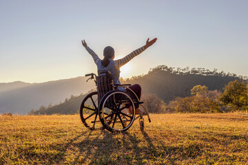 Woman sitting on a wheelchair enjoying sundown on mountain background. - obrazy, fototapety, plakaty