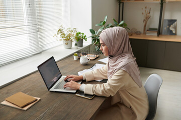 Modern young woman wearing hijab working in office using Internet on laptop, horizontal high angle...