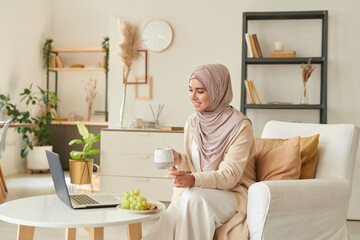 Modern Muslim woman spending free time at home drinking tea and watching comedy film on laptop