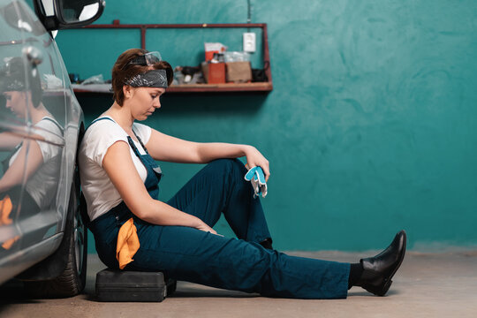 A Tired Sad Caucasian Woman Mechanic Is Sitting Leaning On A Car In An Auto Repair Shop. The Concept Of Women's Work In Male Professions