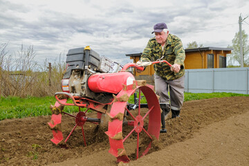 Fototapeta na wymiar An elderly man with a walk-behind tractor forms ridges on the soil for planting potatoes. A farmer cultivates the soil on a cloudy spring day for planting potatoes.