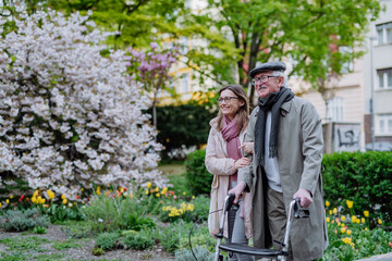 Senior man with walking frame and adult daughter outdoors on a walk in park.