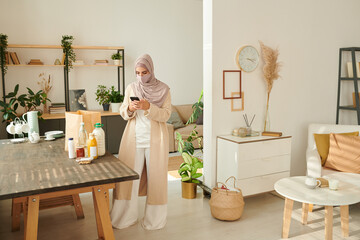 Stylish Muslim woman wearing hijab and mask standing in living room checking something on her smartphone, copy space
