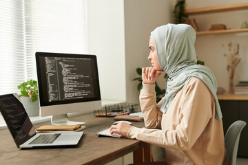 Successful Muslim programmer wearing hijab sitting at desk working on new software using desktop PC and laptop, copy space