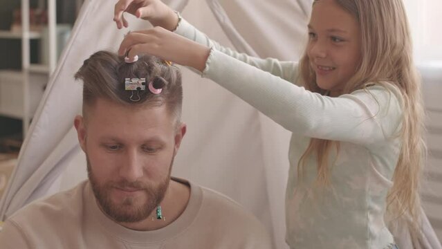Medium slowmo portrait of young modern Caucasian man with beard smiling at camera while his pretty little daughter making ponytails on his hair using paper clips and scrunchies
