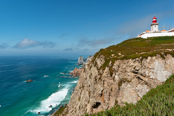 Cliff view of Cabo da Roca, the westernmost point of Portugal, the Iberian Peninsula and Europe and its lighthouse. Calm waves and sunny summer day.