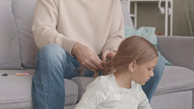 Slowmo of young father with beard sitting on couch in living room while making hair braid for her daughter drawing postcard on Fathers day to him