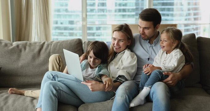 Cute siblings and young parents relaxing on cozy couch with laptop, watching family movie through digital streaming services, spend leisure at modern home using internet and wireless tech, fun concept