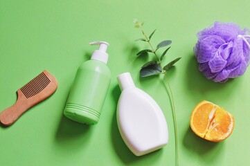 Top view photo natural spa cosmetic products for hair and body care on a green background. White shampoo bottle, liquid soap package, wooden comb, purple sponge, eucalyptus branch and orange