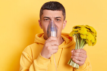Unhealthy man wearing casual style hoodie posing isolated over yellow background, holding bouquet...