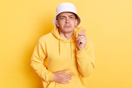 Portrait Of Sick Ill Man Wearing Casual Style Hoodie And Panama Posing Isolated Over Yellow Background, Holding Cookies And Touching His Painful Belly, Suffering Food Poisoning, Overeating.