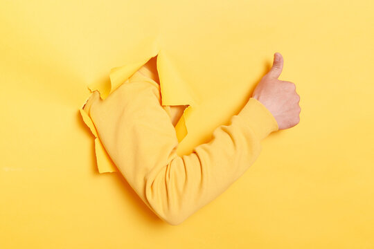 Indoor shot of human hand breaks arm through paper and showing a thumb up sign through a ripped hole, like gesture, isolated over yellow paper background.