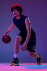 Studio shot of young muscular basketball street player dribbling ball isolated on purple background...