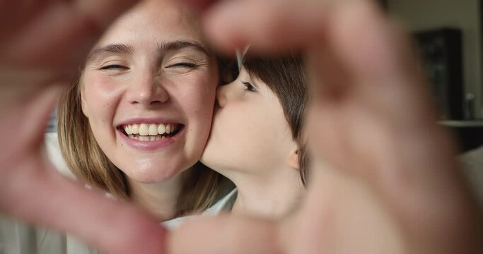 Close up happy faces of young woman her cute little 6s son look at camera through joined fingers showing heart symbol, webcam view, boy kiss moms cheek feeling affection and attachment. Love concept