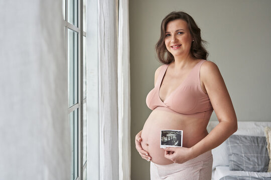 young beautiful pregnant woman holds her hands on her belly in the bedroom and holds an ultrasound picture near her belly. tender photo. concept of future motherhood