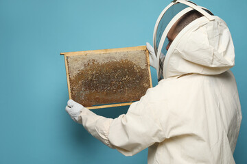 Fototapeta na wymiar Beekeeper in uniform holding hive frame with honeycomb on light blue background, back view