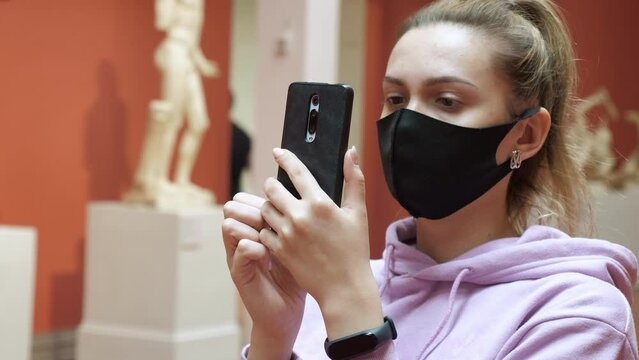 Blonde girl in mask takes pictures of statues she likes in museum on smartphone. Young woman visits exhibition, cultural event keeping protection measures against coronavirus infection