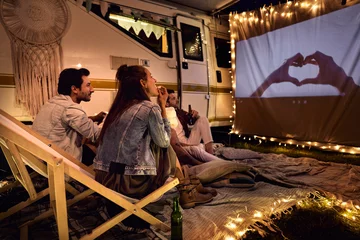 Fotobehang Group of young cheerful people watching a movie on camping site © gpointstudio