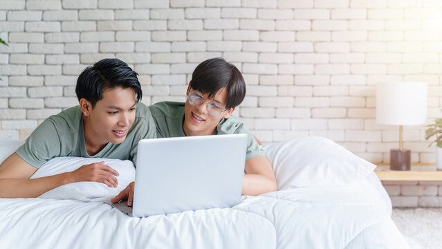 Happy gay asian couple lying on a bed using laptop computer.