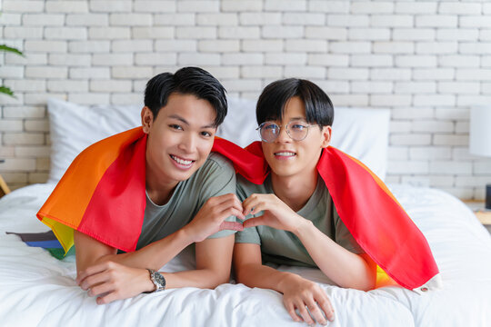 Happy gay asian couple lying on a bed and cover themselves with pride flag. Lovers doing heart-shaped sign with thier hands.