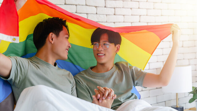 Happy gay asian couple lying on a bed looking at each other and holding pride flag behind their back.