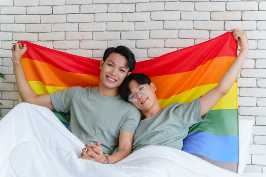 Happy gay asian couple lying on a bed holding hands and pride flag behind their back.