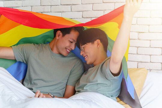 Happy gay asian couple lying on a bed and cover themselves with pride flag.