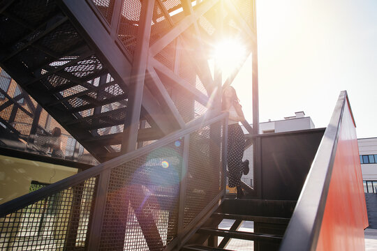 Sunlit image of a woman going upstairs on a heavy metal fire exit stairs. Her gait is light and carefree.