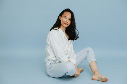 Casual beautiful vitiligo brunette sitting barefoot, presenting deseased patchy part of her face over blue background. Looking peacefully at the camera.