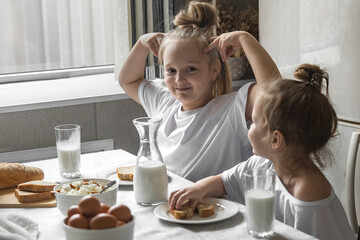 Obraz na płótnie Canvas two sister girls in white t-shirts have breakfast at home in the kitchen with natural and healthy products, cottage cheese and cornbread, milk and boiled chicken eggs