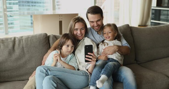 Beautiful young couple and children relax on couch in modern living room having fun using smartphone, watch videos, take selfie pictures, enjoy new mobile application. Tech, leisure with kids concept